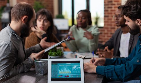 Tablet computer having company turnover on screen standing on table in startup office. Millennial multiethnic businesspeople discussing marketing strategy working at business collaboration